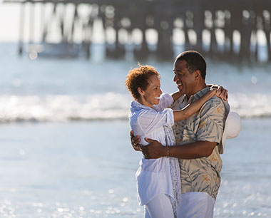 retired couple at the beach with a pier behind them