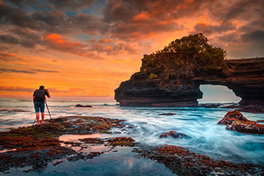 Photograpgher takig picture of sunset over ocean with arch rock